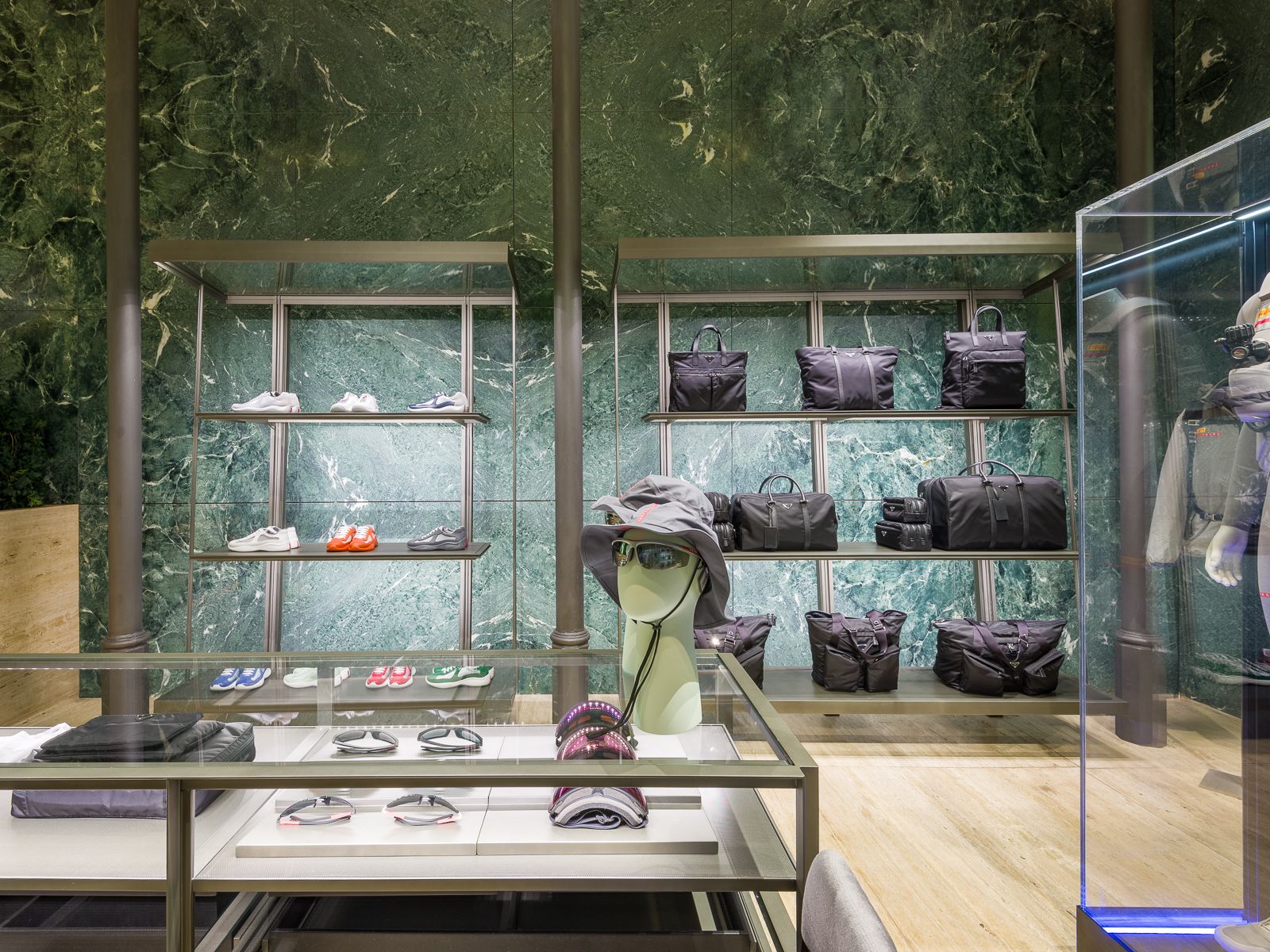 Luxury boutique with green marble walls, showcasing designer sneakers, black bags, and accessories on sleek metal shelving. A glass table displays eyewear and hats, while a mannequin in a performance outfit is encased in a glass display.