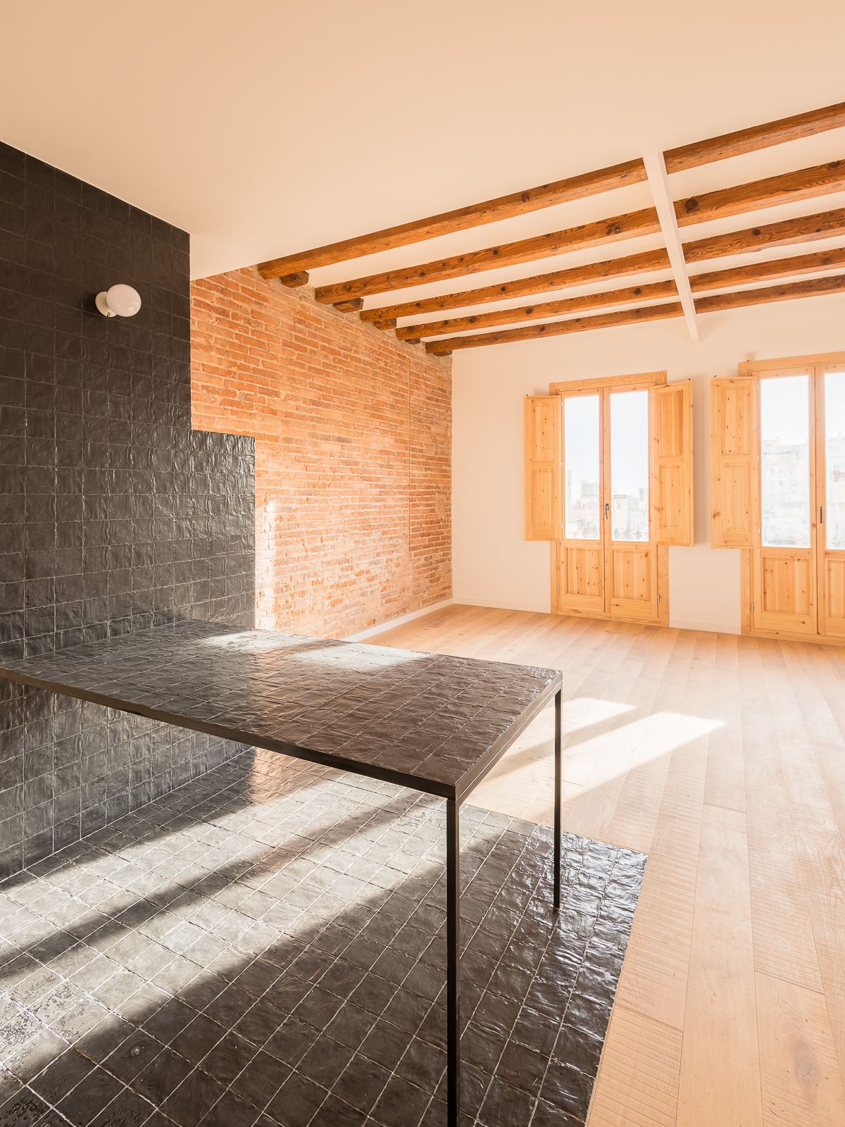Open-plan living area with black tiled countertop, exposed brick wall, wooden ceiling beams, and natural sunlight coming through two large balconies with wooden shutters.