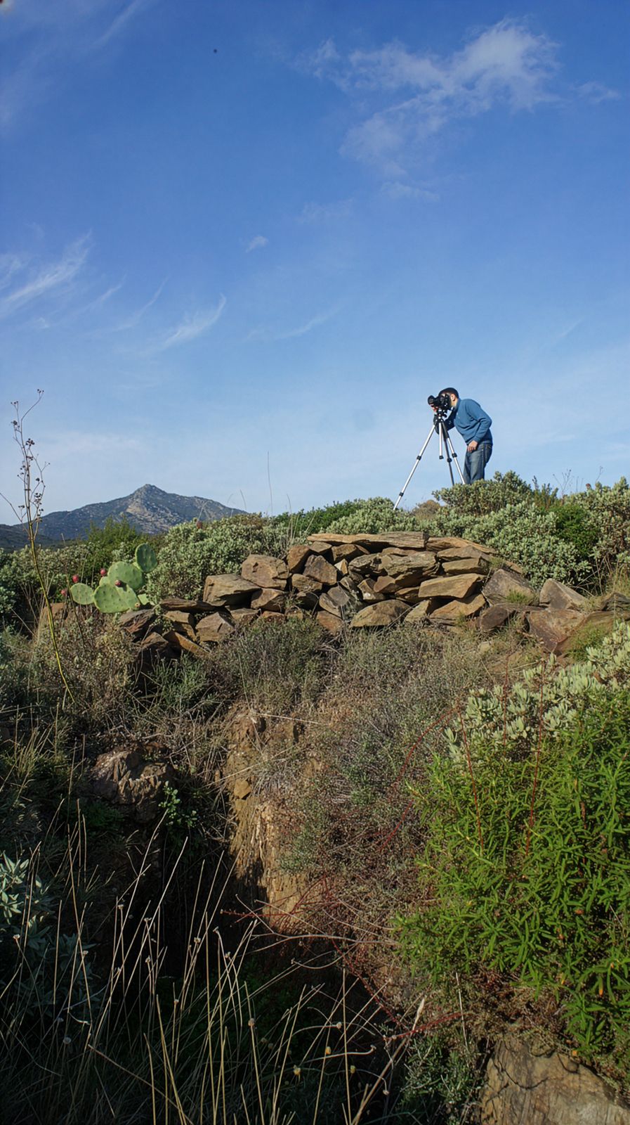Un fotògraf utilitzant un trípode en un turó rocós, capturant una escena de paisatge amb muntanyes al fons sota un cel blau clar.