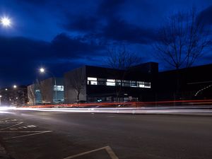 Nighttime street scene with a modern building in the background, car light trails, and streetlights illuminating the area.