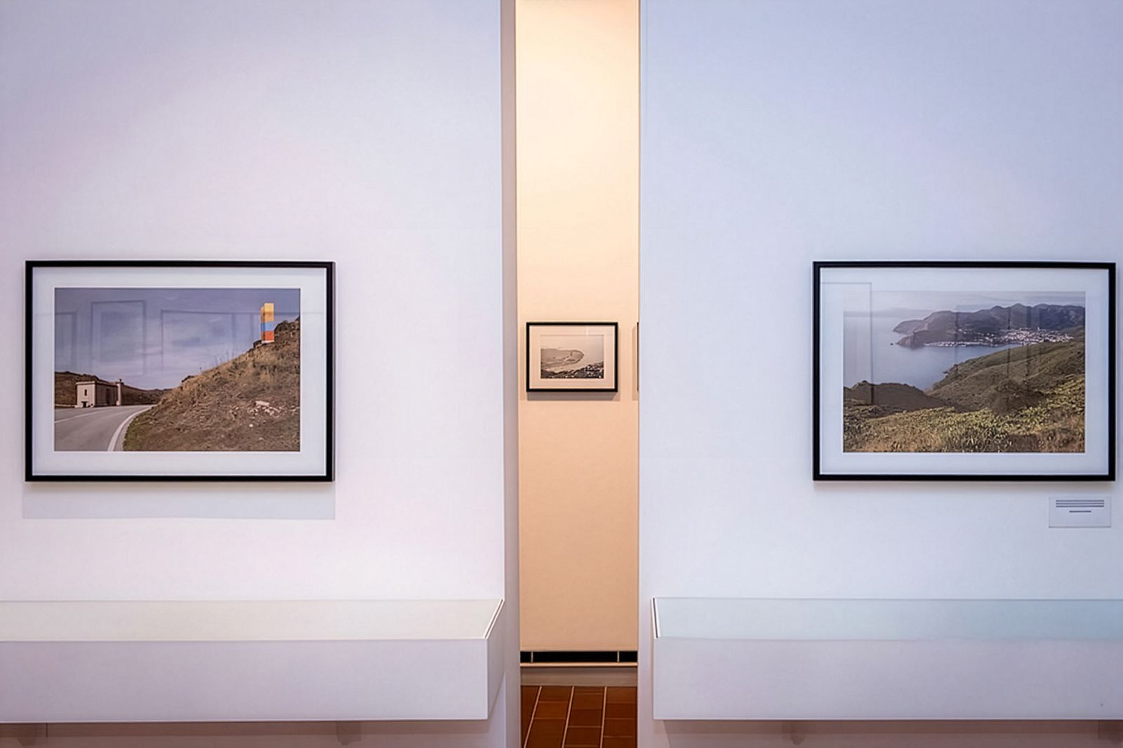 Dues fotografies de paisatge emmarcades exposades en una paret blanca amb una petita fotografia en blanc i negre visible al fons a través d'una obertura.