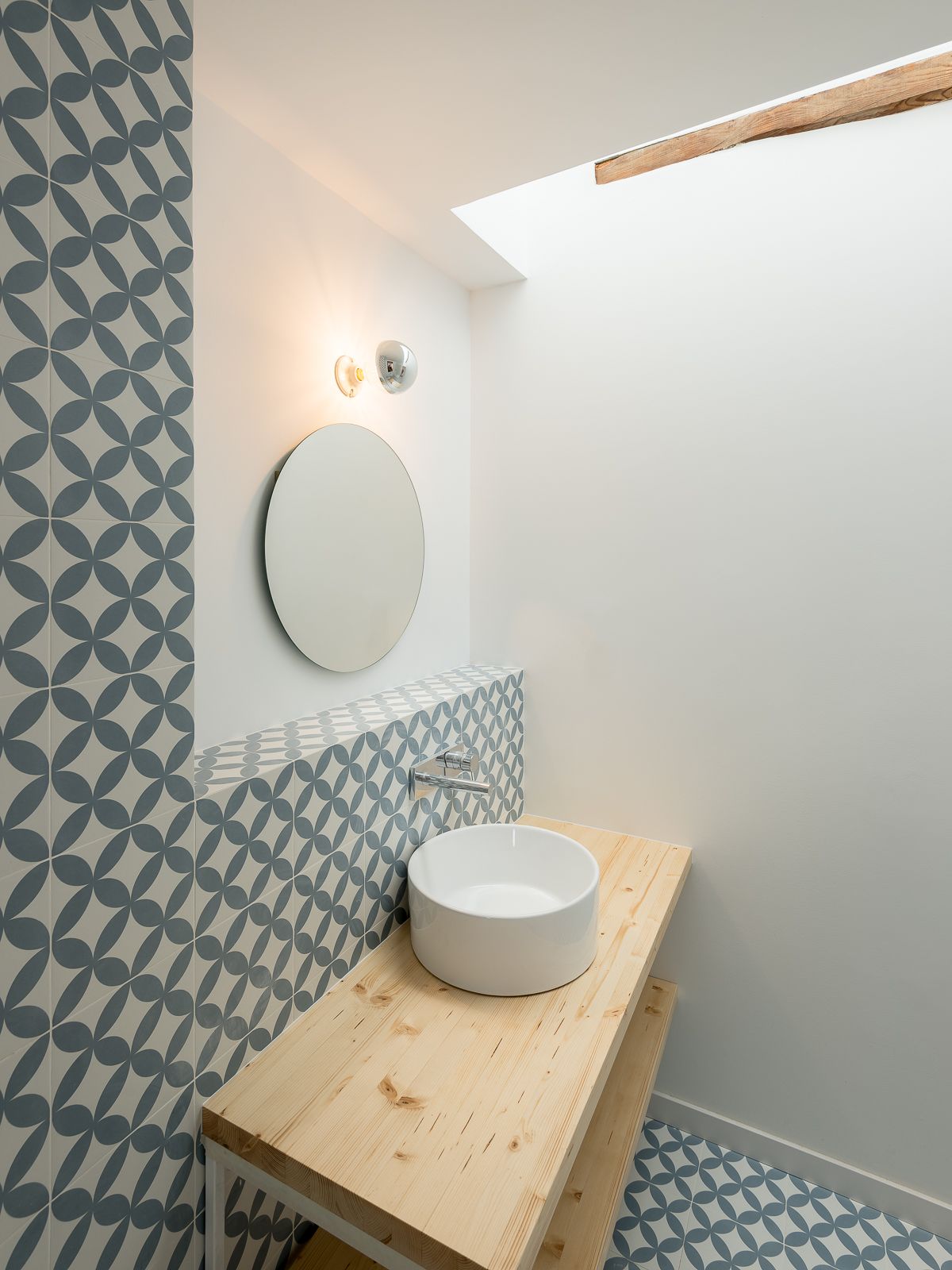 Bathroom with round mirror, vessel sink, wooden vanity, and blue and white geometric tiles, illuminated by natural light from a skylight.