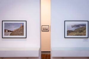 Two framed landscape photographs are displayed on a white wall with a small black and white photograph visible in the background through an opening.
