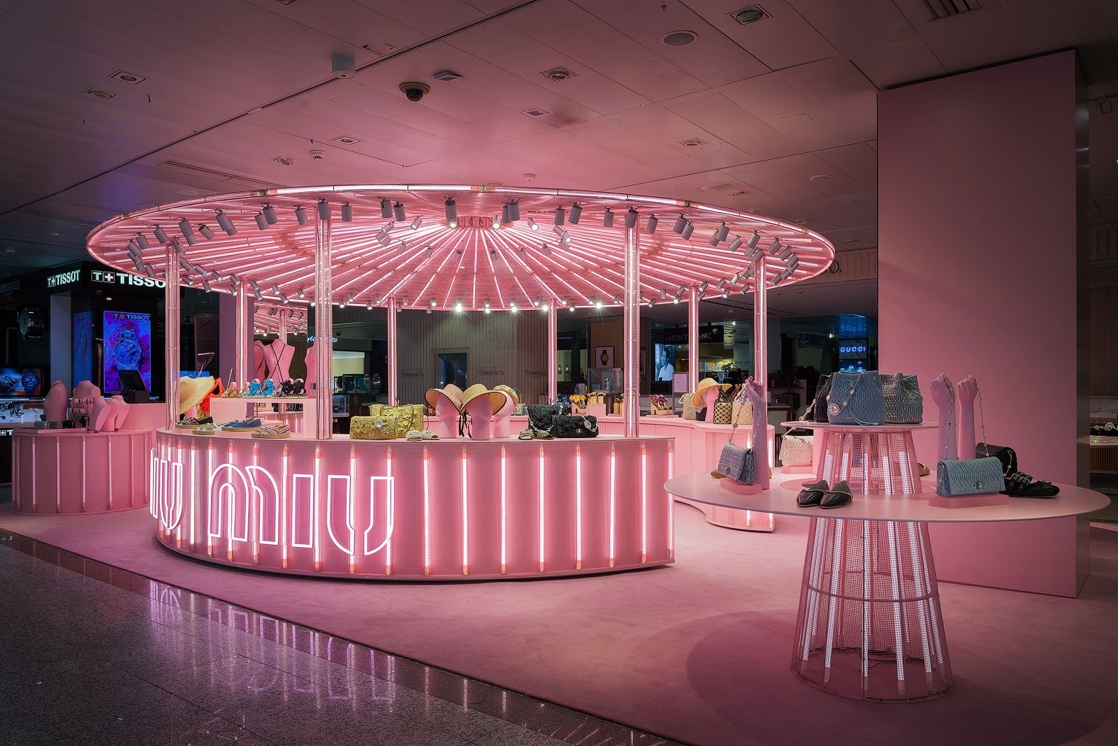 Pink-themed Miu Miu pop-up store with a circular display area, illuminated by vibrant pink neon lights, showcasing various handbags and accessories.