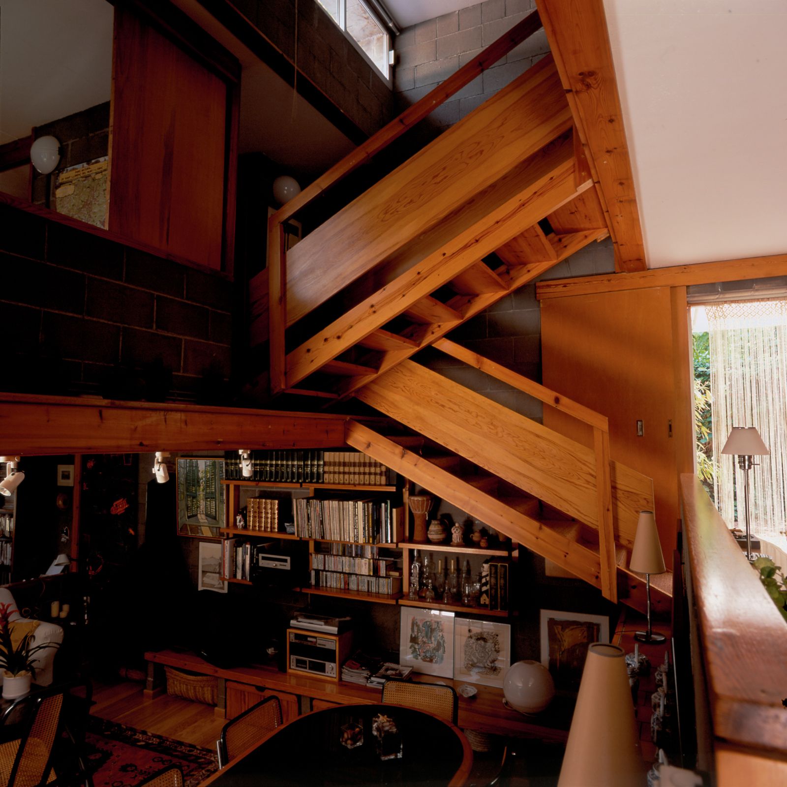 Vista interior amb una escala de fusta, una zona de menjador amb cadires, prestatges plens de llibres i objectes decoratius, i un ambient acollidor creat per la combinació d'elements de fusta i parets de blocs de formigó.
