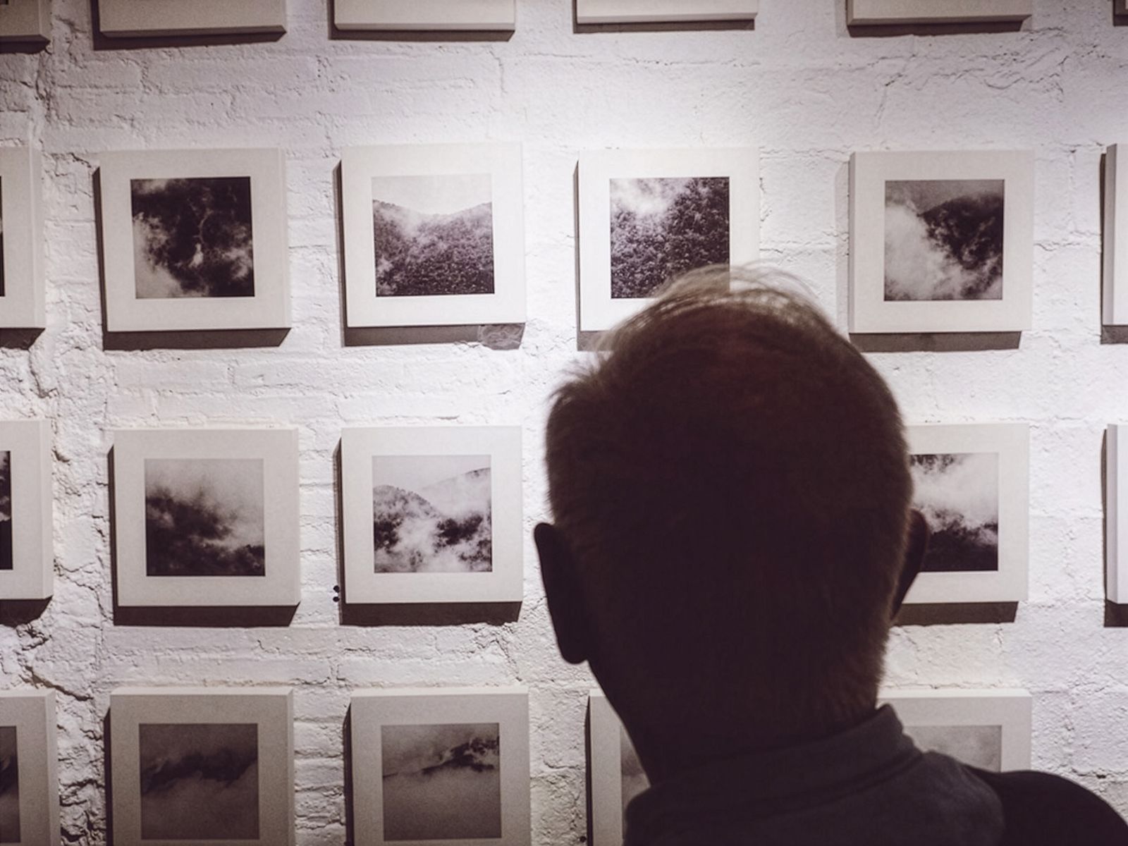 Una persona viendo una gran colección de fotografías cuadradas en blanco y negro de nubes y niebla en una pared de ladrillo blanco.