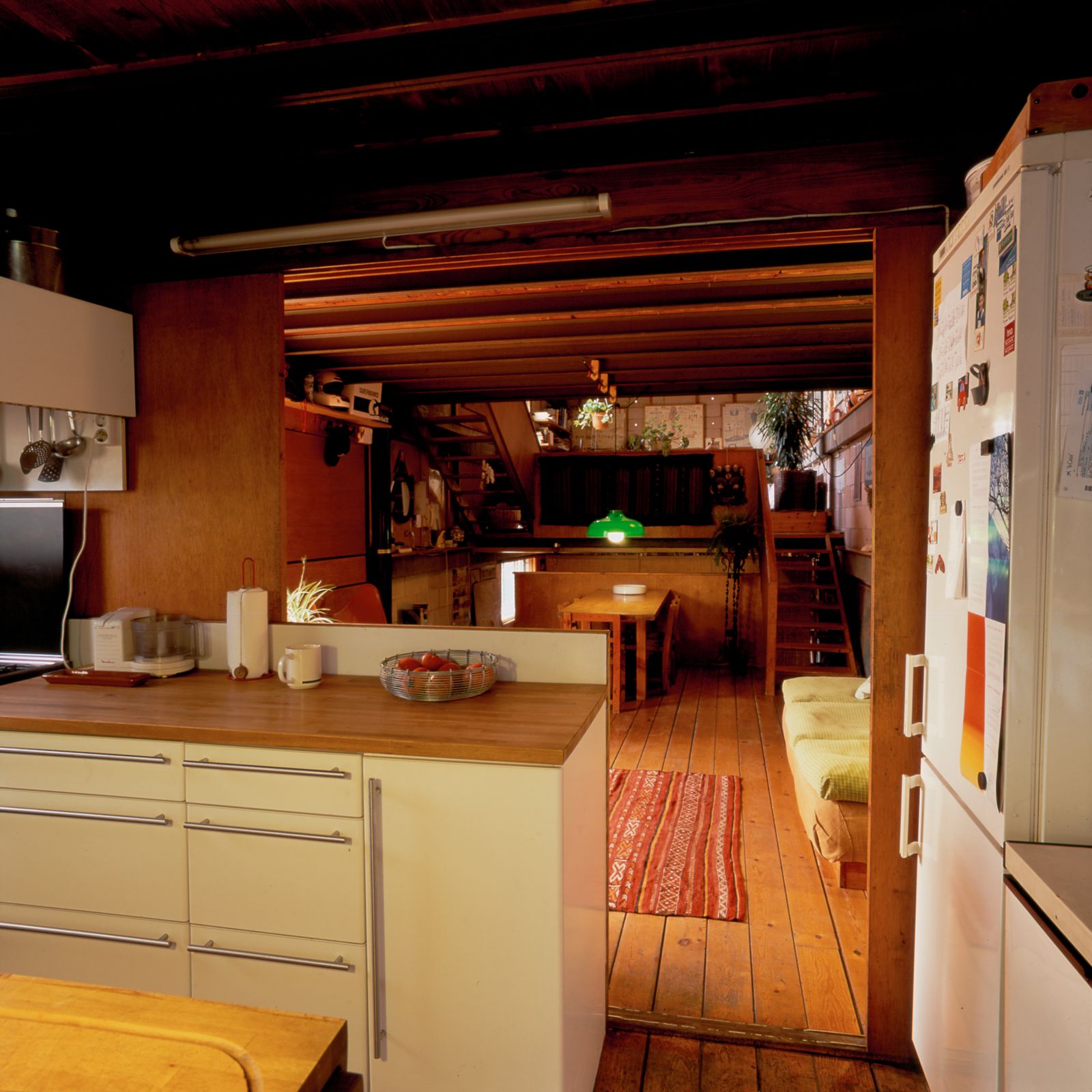 Cozy kitchen with a wooden floor and ceiling, white cabinets, leading to a dining area with a colorful rug, a wooden table, green pendant light, and an open air wooden staircase in the background.