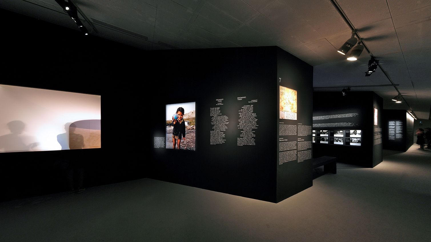 Wide-angle view of a modern museum exhibition space featuring matte black display walls with text blocks, a lit photograph of a child, a glowing projection screen on the left, and a gallery pathway continuing into the background.