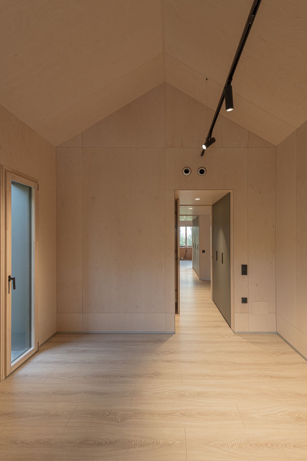 Vista interior d'una habitació amb un sostre inclinat, panells de fusta clara, il·luminació de carril i una porta que condueix a un passadís, amb una finestra a l'extrem llunyà.