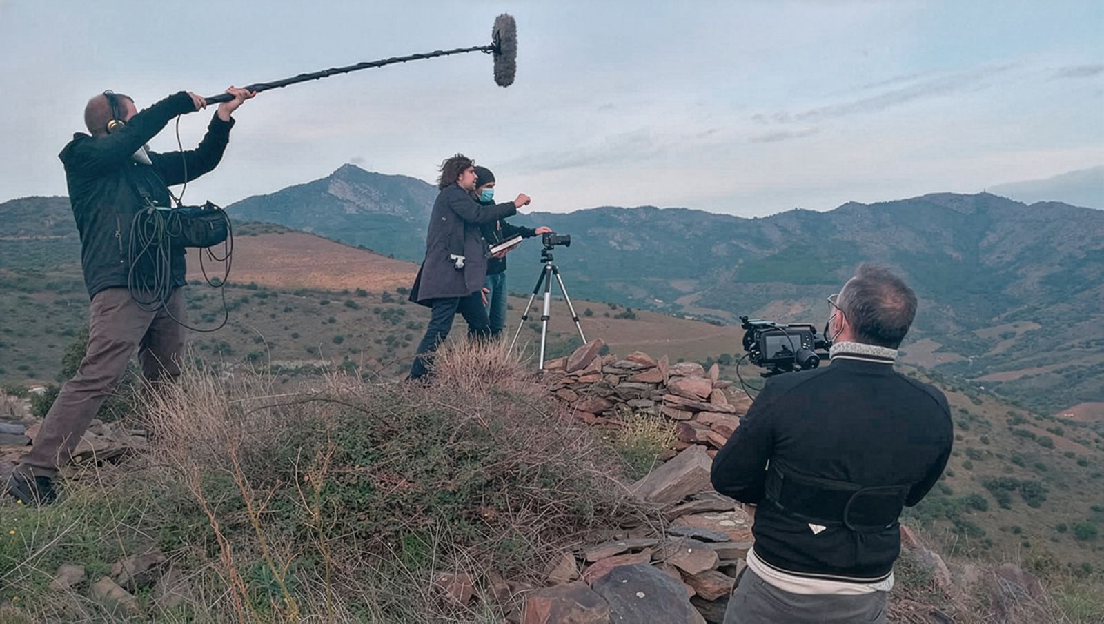Un equip de filmació rodant en un turó amb un teló de fons muntanyós escènic. L'equip inclou un càmera i una persona sostenint un micròfon de perxa, estan filmant dues persones que munten un trípode amb una càmera.