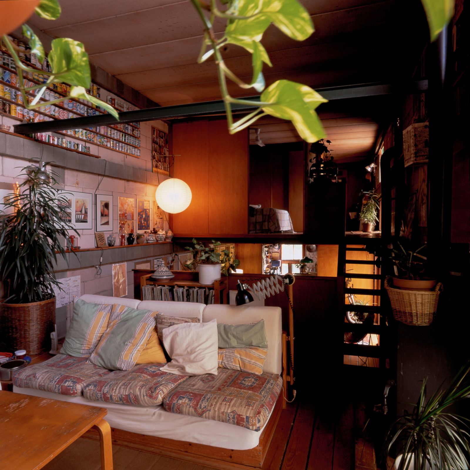 Cozy living room with a colorful sofa, wooden furniture, indoor plants, and a white pendant light, featuring a staircase and a wall decorated with art and collectibles.