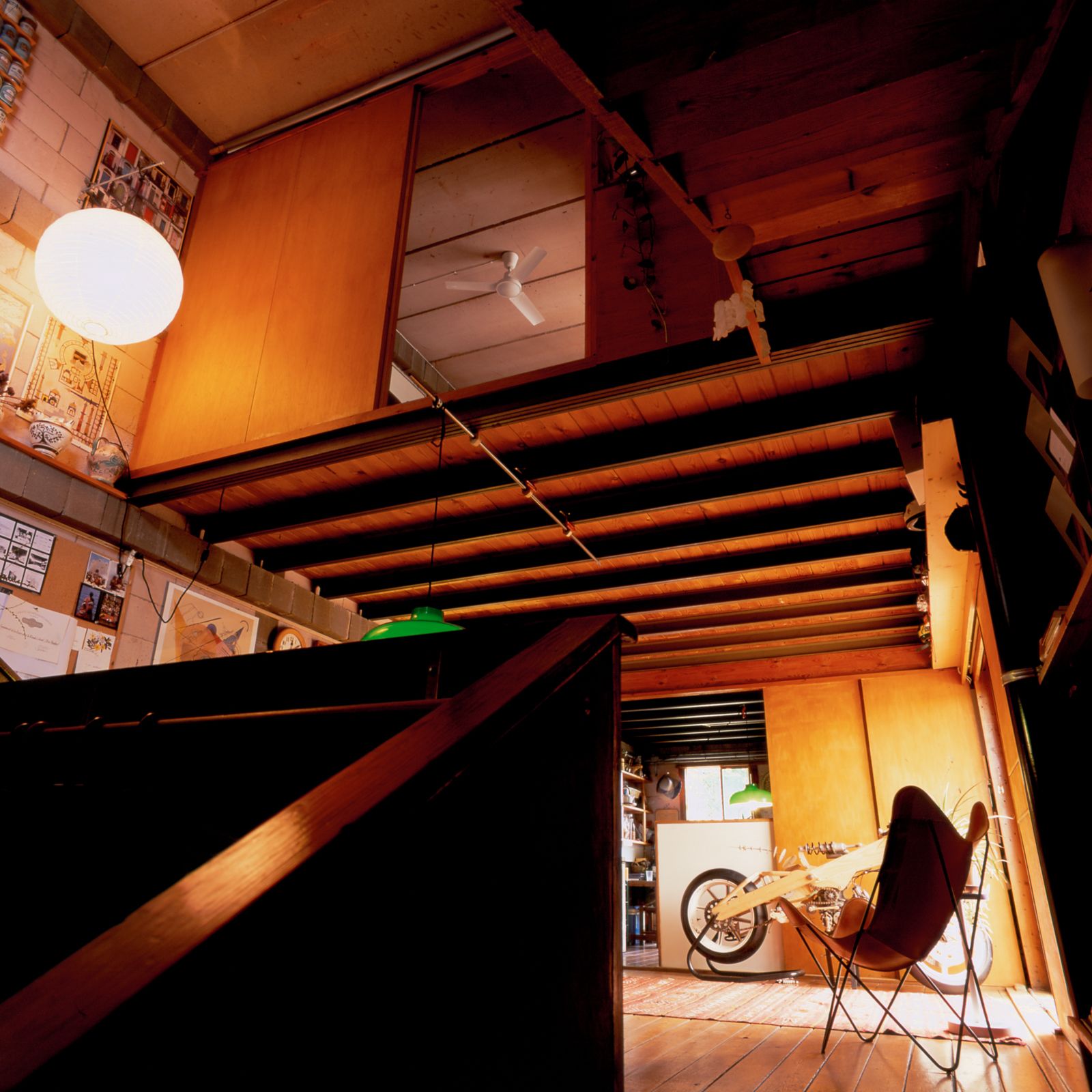 Low-angle interior view of a warm, eclectic space with exposed steel beams, sliding wooden panels opening into a loft area with a ceiling fan. In the ground floor, a wood motorcycle prototype is visible in the background.