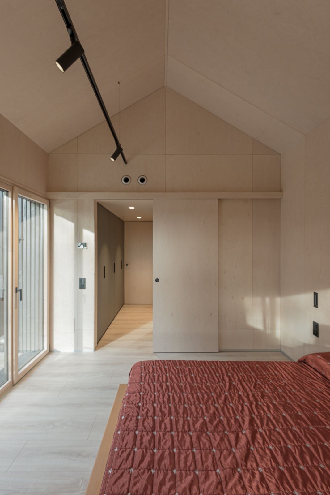 Habitació amb un sostre inclinat alt, amb un cobrellits vermell, grans portes de vidre i parets i terra de fusta minimalistes.