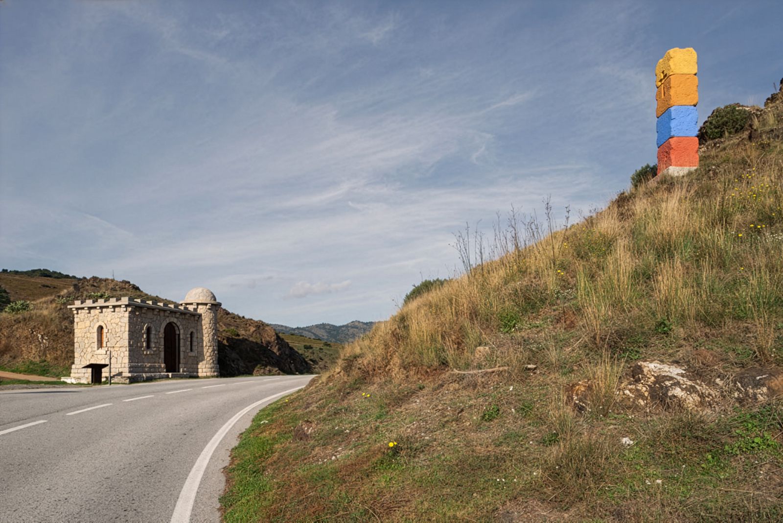 Una carretera sinuosa amb un edifici de pedra minúscul que sembla un castell a l'esquerra i un pilar apilat de colors a la dreta, amb turons ondulants al fons sota un cel blau.